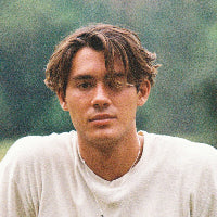 Man wearing a white t-shirt and dark pants standing outdoors with greenery in the background