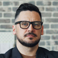 Man wearing glasses and a black shirt in front of a brick wall with decor items.